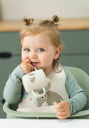 Kind in einem Hochstuhl trägt eine hellgraue Lätzchen mit Tiermotiven, hält einen cremefarbenen Becher mit kleinem Tiermotiv in der Hand.
