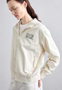 Casaco corta-vento de cor creme feito de material leve. Apresenta um fecho com zip, punhos elásticos e um logótipo EA7 bordado no peito.