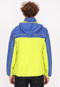 Chaqueta cortavientos con un cuerpo amarillo brillante y mangas azules, con capucha, costuras en contraste y una textura ligera y suave.