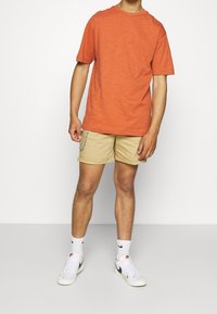 Orange strukturiertes T-Shirt, beige Shorts mit Ketten-Detail, weiße Nike Sneaker und weiße Socken mit schwarzem Logo, stehend vor einem neutralen Hintergrund.