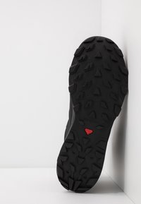 Schwarze Sportschuhsohle mit tiefen, kantigen Profilen für besseren Grip, ausgestattet mit einem roten Akzentlogo nahe der Ferse zur Identifikation.