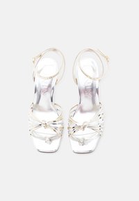 Ασημένια σανδάλια με ψηλό τακούνι και λουράκια στον αστράγαλο με κόμπο στη μύτη, εμφανιζόμενα από πάνω σε λευκό φόντο.