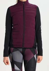 Gilet senza maniche imbottito di colore viola con zip frontale, indossato sopra una maglietta nera a maniche lunghe e leggings a fantasia geometrica in nero, viola e grigio.