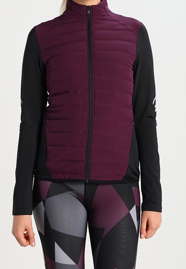 Gilet senza maniche imbottito di colore viola con zip frontale, indossato sopra una maglietta nera a maniche lunghe e leggings a fantasia geometrica in nero, viola e grigio.