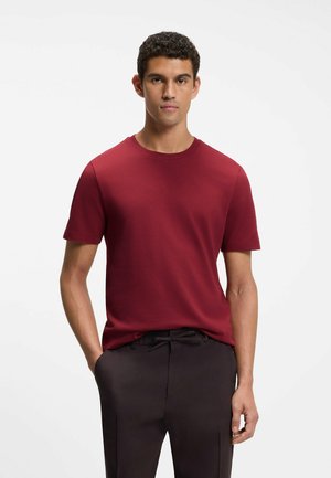 Man wearing a short-sleeve textured red shirt and black pants, standing with one hand in pocket against white background.