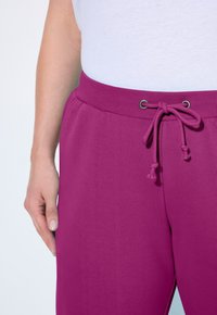 Pantaloni jogger aderenti in tessuto rosa brillante con vita elastica, chiusura con coulisse e tasche laterali, caratterizzati da una texture liscia.