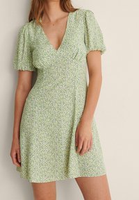 Robe florale verte avec des manches bouffantes, un décolleté en V et un ourlet évasé, ornée de petits motifs floraux violets et bleus sur un fond clair.
