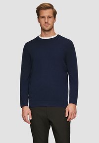 Marineblauwe gebreide crewneck trui van zacht materiaal met lange mouwen en ribgebreide zoom, gedragen met zwarte broek. Gladde textuur, minimaal ontwerp.