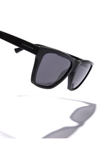 Ochelari de soare din plastic negru cu un design unghiular și lentile gri. Prezintă un logo pe braț. Umbră vizibilă pe un fundal deschis.