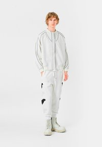 Chaqueta bomber blanca con ribetes negros, combinada con joggers blancos con acentos bordados en negro y botas de combate color crema. Diseño minimalista.