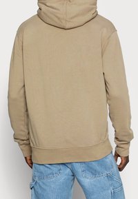 Beige väritetty huppari rentolla istuvuudella, resorikauluksella ja helmalla. Pehmeä pinta, saumaton muotoilu, eikä näkyvää merkintää tai grafiikkaa.