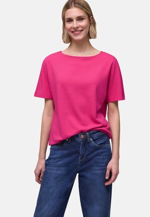 Smilende kvinde med kort blondt hår iført en struktureret, lyserød kortærmet top og blå jeans mod en ensfarvet baggrund.