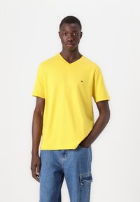 Yellow V-neck t-shirt made of cotton, featuring short sleeves and a small logo on the left chest, paired with blue cargo jeans.