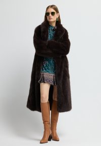 Abrigo largo de piel sintética marrón oscuro, vestido de patrón verde azulado con dobladillo con volantes, y botas de cuero marrón hasta la rodilla con tacón ancho y detalle de cremallera.