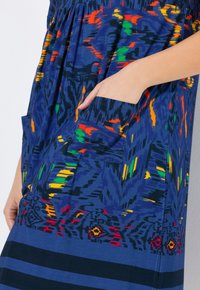 Granatowa sukienka z żywym, wielokolorowym abstrakcyjnym wzorem. Posiada plisowany krój i kieszeń z przodu. Gładki materiał.