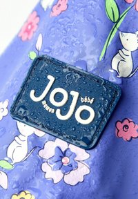 Purplos, vandeniui atsparus audinys su gėlių raštais rožinėmis, mėlynomis ir žaliosiomis spalvomis. Yra mėlynas etiketas su užrašu "Jojo bébé" iškilusiomis baltosiomis raidėmis.