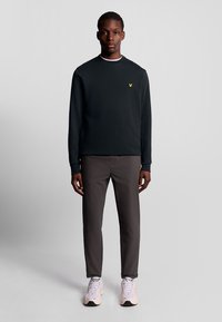 Zwart sweatshirt met ronde hals, gele logo op de borst, gecombineerd met grijze broek en lichte sneakers, staand tegen een effen achtergrond.