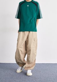 Person som bär en teal sporttröja med vita ränder, beige baggy cargo-byxor och vita sneakers med svarta ränder, stående på en blå matta.