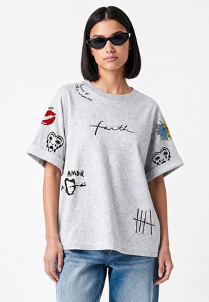Donna che indossa occhiali da sole neri, una T-shirt oversize grigio chiaro con stampe grafiche nere e rosse e jeans blu su sfondo bianco.