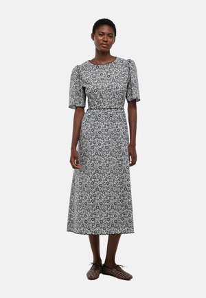 Femme debout portant une robe mi-longue à manches courtes, avec une taille cintrée et un motif floral noir et blanc petit et présent sur toute la surface.