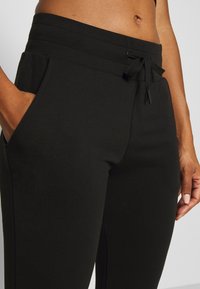 Gros plan d'une personne portant un pantalon noir à cordon avec les mains dans les poches, montrant une vue recadrée du ventre et des bras.