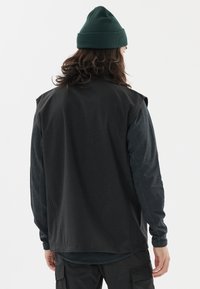 Schwarze ärmellose Jacke über einem blauen Langarmhemd. Verfügt über eine glatte Textur, Schulternaht und einen hohen Kragen. Grüner Beanie auf dem Kopf.