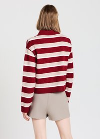 Pull rayé rouge et beige avec un col et un ourlet côtelés, à manches longues. Associé à un short beige, présentant un design court.