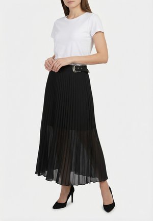 Mujer con una blusa blanca de manga corta, falda larga negra plisada y transparente con cinturón, y tacones altos negros puntiagudos, de pie sobre fondo blanco.