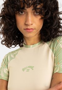 Rash guard beige con estampado botánico verde en las mangas, que presenta un logo en un suave verde en la parte frontal. Textura de tela suave.