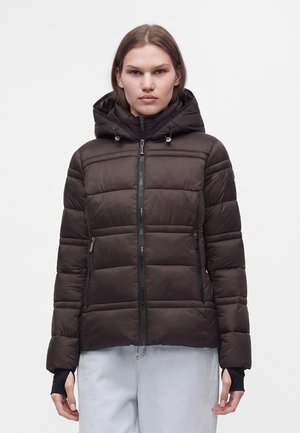Femme portant une doudoune noire à capuche avec une fermeture éclair à l'avant et un pantalon clair, debout sur un fond blanc uni.