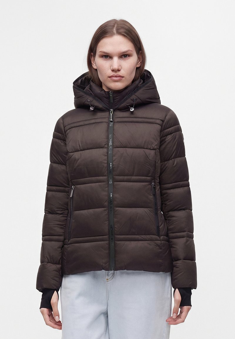 Femme portant une doudoune noire à capuche avec une fermeture éclair à l'avant et un pantalon clair, debout sur un fond blanc uni.