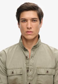 Camisa de botones color caqui con dos bolsillos en el pecho, un cuello alto y detalles de costura visibles, llevada por una persona con pelo corto.