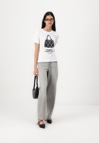 Hvit T-skjorte med svart grafisk design, som viser en håndveske og tekst. Kombinert med løse grå jeans og svarte sko, og holder en sirkulær veske.