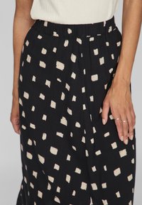 Svart tygkjol med ett spritt mönster av krämfärgade rutor, elastisk midja och lös passform. Handen vilar på kjolens sida.