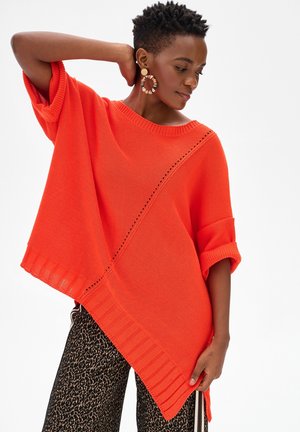 Osoba z krótkimi włosami w pomarańczowym asymetrycznym swetrze i spodniach w panterkę, pozująca z jedną ręką za głową i patrząca w dół.