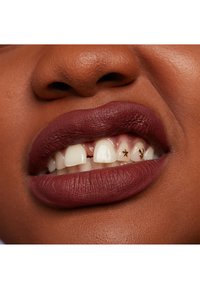 Matte mörkröd läppstift på läpparna, som avslöjar vita tänder med guldstjärnor som accent. Huden har en slät, varm ton med en subtil textur.