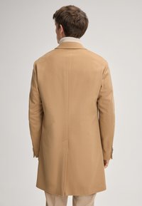 Man in een camel-kleurige jas en lichtbeige coltrui, met zijn rug naar de camera tegen een effen lichtgrijze achtergrond.