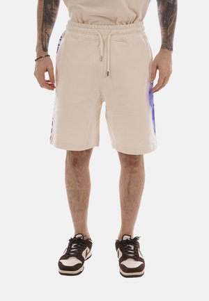 Hombre con pantalones cortos con cordón color crema y paneles laterales morados, y zapatillas negras y blancas, de pie con los brazos relajados.