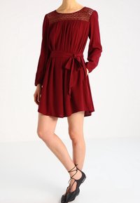 Burgundy mekko pitkillä hihoilla, vyötärösidonta ja pitsiyksityiskohta kaula-aukossa. Väljä kangas, jossa levenevä hame, polvipituinen.