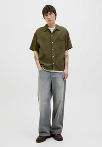 Olijfgroene, kortemouwen, gewatteerde shirt met een enkele borstzak, gecombineerd met lichtblauwe, losse jeans en donkerbruine schoenen.