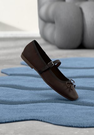 Dark brown Mary Jane flat shoe with buckle and bow detail resting on wavy blue textured rug and gray cushioned sofa in background.