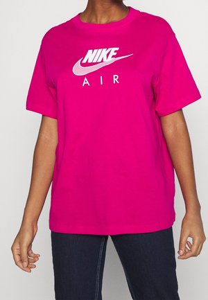 Personne portant un t-shirt Nike Air rose vif avec un logo blanc et un jean foncé, debout devant un fond clair uni.
