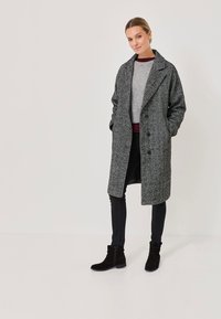 Femme portant un long manteau en chevrons gris, un pull gris, un pantalon skinny noir et des bottines noires, se tenant contre un fond uni.