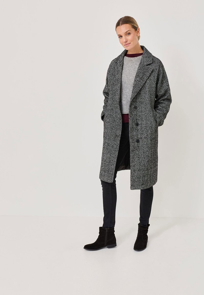 Femme portant un long manteau en chevrons gris, un pull gris, un pantalon skinny noir et des bottines noires, se tenant contre un fond uni.