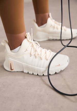 Zbliżenie na stopy w białych butach treningowych Nike obok czarnej skakanki na szarej podłodze.