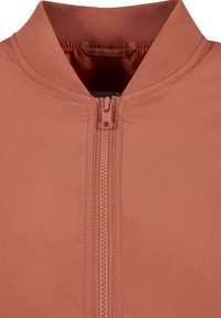 Veste de couleur rouille avec fermeture éclair à l'avant et col côtelé, montrant un gros plan de la texture du tissu et des coutures.