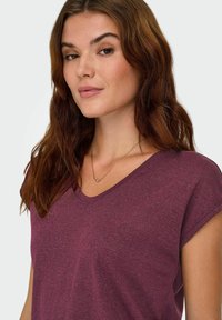 Une femme avec de longs cheveux bruns portant un haut à manches courtes bordeaux et un délicat collier en or avec un petit pendentif rond.