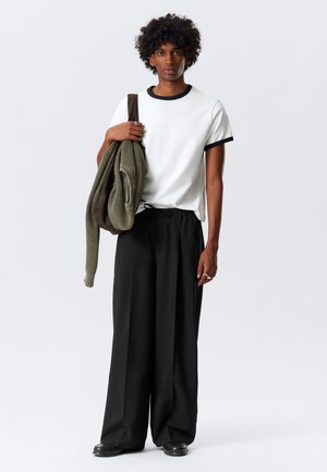 White t-shirt with black ribbed collar, loose black trousers, olive green knit sweater over one arm, and black shoes.