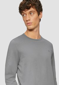 Langärmliges graues Shirt mit rundem Ausschnitt. Weicher Stoff, körpernahe Passform und ein kleines Logo auf der linken Brust. Glatte Stoffoberfläche.