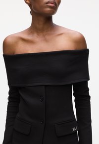 Black off-the-shoulder top with long sleeves, buttons down the front, and pockets featuring embroidered details. Smooth fabric texture.